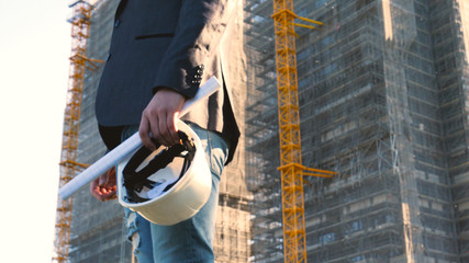 Obraz na płótnie Canvas Portrait of a successful young handsome engineer, architect, builder, businessman, wearing a white helmet, in a suit, holding a project in his hand, a skyscraper background and a construction site.