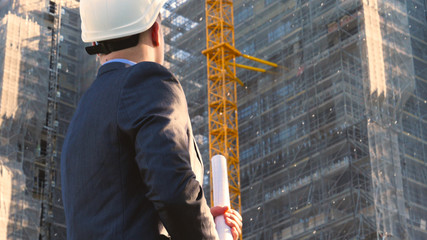 Portrait of a successful young handsome engineer, architect, builder, businessman, wearing a white helmet, in a suit, holding a project in his hand, a skyscraper background and a construction site.