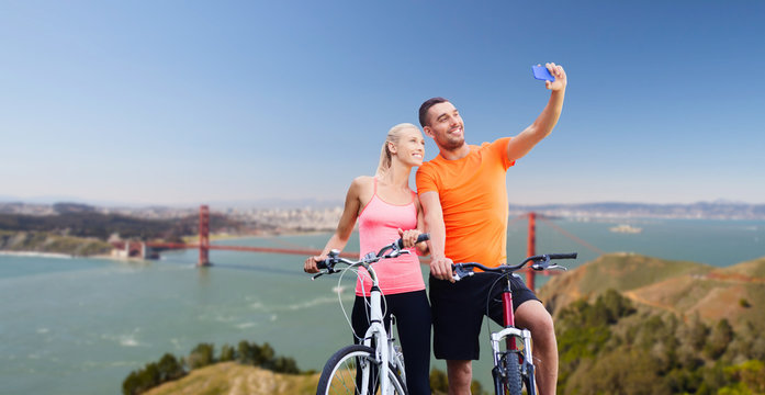fitness, sport, technology and healthy lifestyle concept - happy couple riding bicycles and taking selfie by smartphone over golden gate bridge in san francisco bay background
