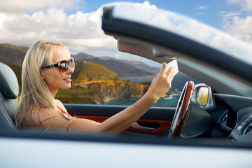 travel, road trip and people concept - happy young woman in convertible car taking selfie by...