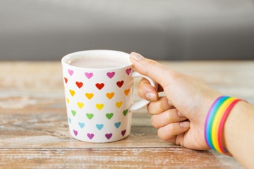 homosexual and lgbt concept - close up of female hand with cacao drink in cup with heart pattern and gay pride awareness wristband on wooden table