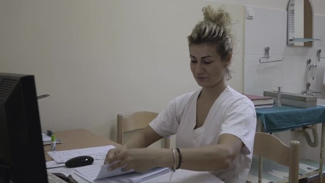 Portrait of pretty blonde nurse in white uniform while sitting and working with patient documentation in ambulatory room at hospital, young woman enters data from papers in the computer, close up