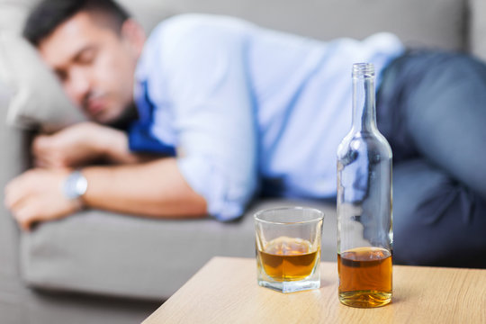 Alcoholism, Alcohol Addiction And People Concept - Bottle With Glass Of Whiskey On Table And Sleeping Drunk Man