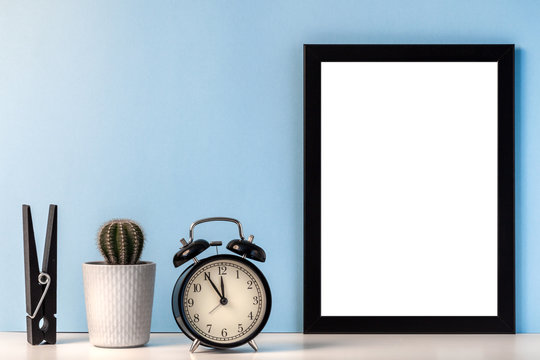 Mockup with a black empty frame with a white center against the background of a blue wall with a black alarm clock, cactus and oversizde clothes peg
