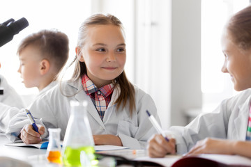 education, science and children concept - kids studying chemistry at school laboratory and writing to workbooks