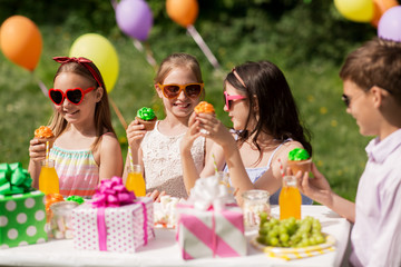 holidays, childhood and celebration concept - happy kids in sunglasses sitting at table on birthday party at summer garden and eating cupcakes