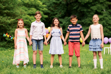 holidays, childhood and celebration concept - happy kids holding hands on birthday party at summer park