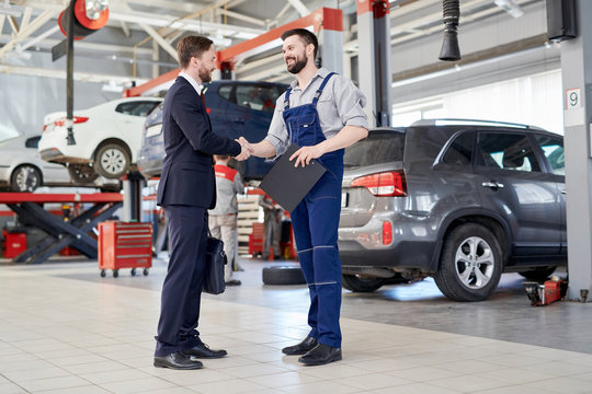 Full length portrait of handsome businessman shaking hands with worksman while standing in production workshop of modern car factory, copy space