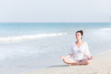 Fototapeta na wymiar Young pregnant woman practicing yoga at sea at summer vacation, sitting in the lotus position on the beach with white sand. Healthy pregnancy lifestyle concept