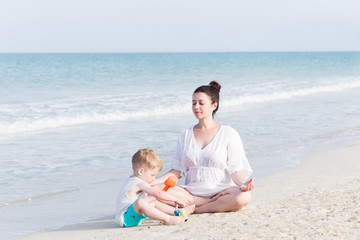 Portrait of happy pregnant mother relax near playing son at sea beach with white sand. Happy family at summer vacation concept