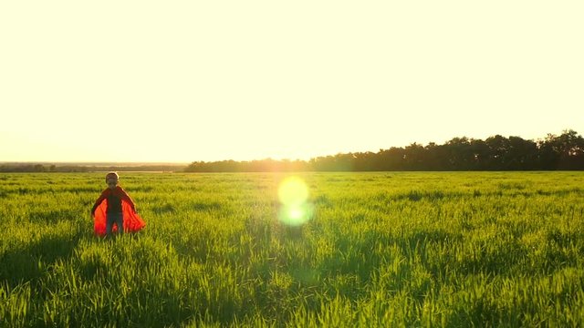 The child in the costume of a superhero in a red cloak is standing in the green lawn against the backdrop of the sunset.