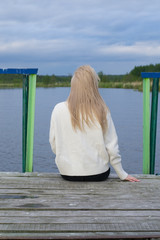 Sad young woman is sitting on pier by lake 