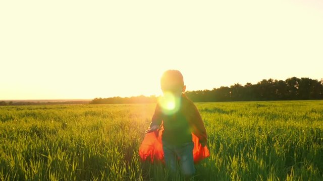 A child in the costume of a superhero in a red cloak runs across the green lawn against the backdrop of a sunset toward the camera