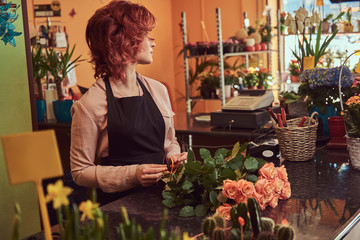 Charming redhead female florist wearing uniform making a beautiful flower composition while standing at the counter of flower shop.