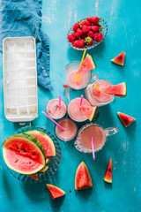 Watermelon an strawberry agua fresca cocktail , summer fresh drink on different glass container over a blue  background and with fruits pieces around, ice container an colored straws.