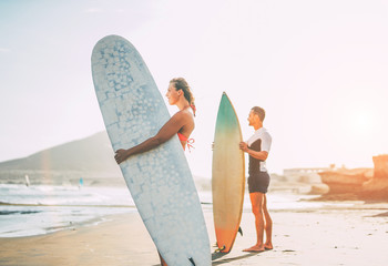Young couple of surfers standing on the beach with surfboards preparing to surf on high waves during a magnificent sunset - People, lifestyle, sport concept - Focus on woman