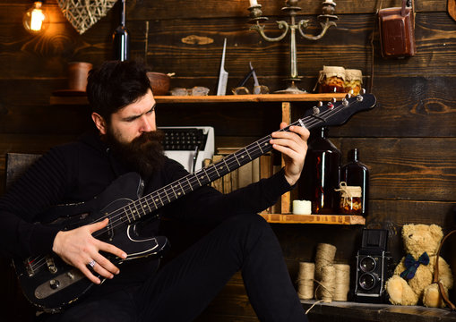 Best weekend. Man bearded musician enjoy evening with bass guitar, wooden background. Man with beard holds black electric guitar. Guy in cozy warm atmosphere play relaxing soul music