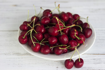 Ripe cherry berries with water droplets on a white plate on a white wooden background, clean, healthy and tasty fruit.