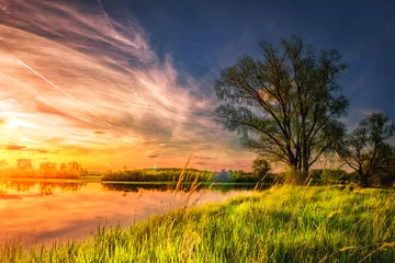 Foto op Plexiglas geweldig landschap van de zomer natuur op de oever van de rivier bij zonsondergang met kleurrijke bewolkte hemel. Perfecte scène grote boom op met gras begroeide oever van het meer. Gras gloeien op warm zonlicht in de avond. © dzmitrock87