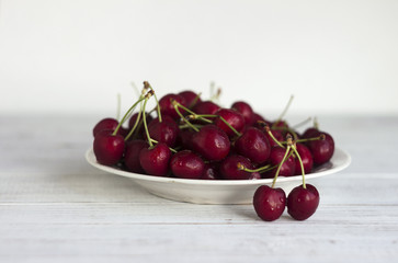 Naklejka na ściany i meble Ripe cherry berries with water droplets on a white plate on a white wooden background, clean, healthy and tasty fruit.