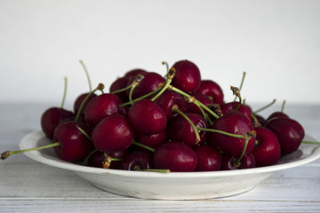 Ripe cherry berries with water droplets on a white plate on a white wooden background, clean, healthy and tasty fruit.