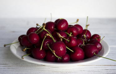Ripe cherry berries on a white plate on a white wooden background, useful and tasty fruit.
