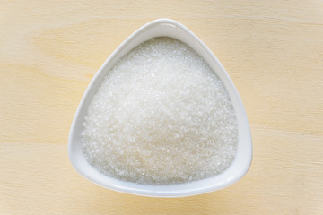 Fototapeta na wymiar Danger of fat or obesity and risk of diabetes mellitus concept : Top view of pure white refined, bleached sugar crystal / fine granulated sugar sweetener in a triangular ceramic bowl on a wood table.