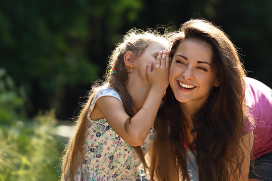 Happy Kid Girl Whispering The Secret To Her Laughing Young Mother In Ear With Fun Face On Summer Green Tree And Grass Background.