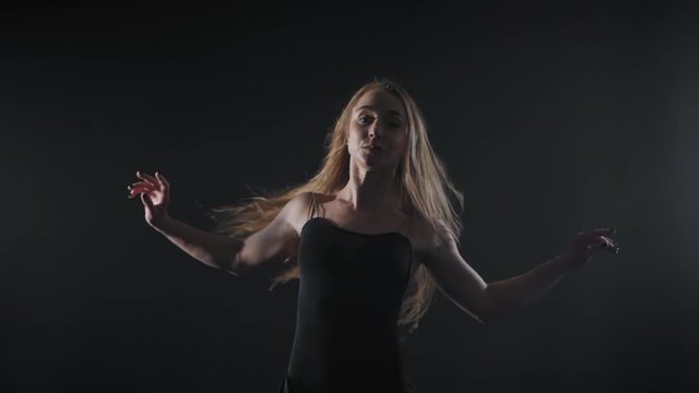 Professional ballerina dancing ballet in spotlights smoke on big stage. Beautiful caucasian young girl with long hair wearing black tight dress on monochrome background.