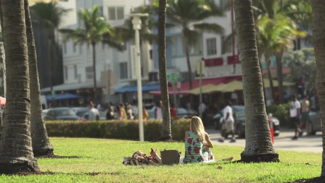Miami beach cityscape, street view. Young girl sitting in the Lummus park at Ocean drive, South Beach - April 2018: Miami beach, Florida, US
