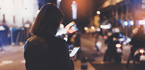 Girl pointing finger on screen smartphone on background bokeh light in night city street, blogger hipster girl using in hands mobile phone, lights auto taxi; lifestyle online wifi internet concept