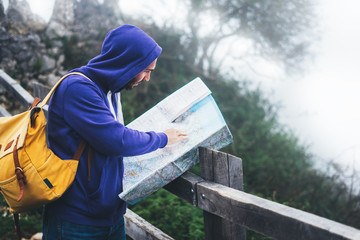 Hipster young man with backpack holding in hands and looking on map spain of foggy mountain, tourist traveler hiker on background valley landscape panoramic view, trip in northern spain basque country