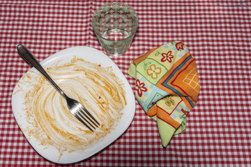 meal plate finished with remains of tomato sauce on a tablecloth with a napkin and glass