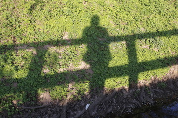 Mann oder Frau steht auf einer Brücke, ihr Schatten fällt auf eine Wiese am Ufer des Baches