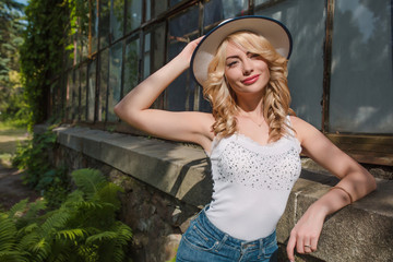 Smiling young lady wearing hat and trendy outfit posing near the greenhouse. Space for text