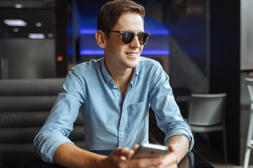 Happy young man in glasses, with phone in hand, sitting in cafe, suitable for advertising, text insertion