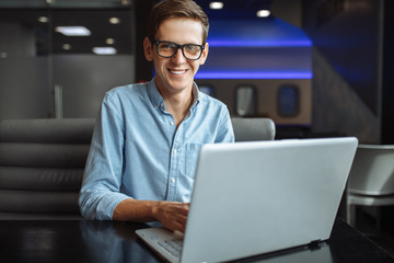 Portrait of a young man with a good mood, a businessman in a shirt and glasses, who works on a laptop in a cafe, can be used for advertising, text insertion.