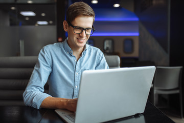 Portrait of a young man with a good mood, a businessman in a shirt and glasses, who works on a laptop in a cafe, can be used for advertising, text insertion.
