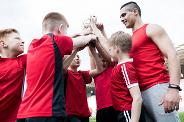 Portrait of happy junior football team holding trophy together and cheering after winning match in...