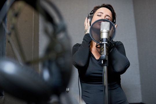 Beautiful woman in black dress wearing headphones and singing with mic keeping eyes closed in studio. 