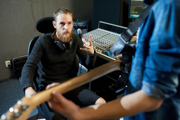 Bearded man sitting at console in recording studio and talking to crop man with guitar preparing...