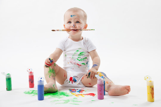 Cute little baby painting with paintbrush and colorful paints on white background. Brush in the mouth.