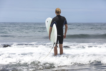 The surf enters the water. Male surfer entering the sea with his board in a black surfing suit. Tenerife, Spain. Surfer entering the ocean .Ready for a great surfing day. - Powered by Adobe