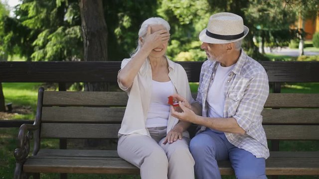 Handsome senior man with gray beard surprising his mature beautiful woman with white hair with marriage proposal while two sitting on a bench. Female with closed eyes getting engagement ring from man