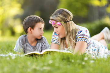Young mother and son lying on grass in park and reading a book