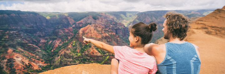 People hiking in Hawaii banner hikers pointing at Kauai mountains. happy hiker couple healthy lifestyle outdoors looking at Waimea canyon view. Young couple in nature in Kauai, Hawaii, USA.