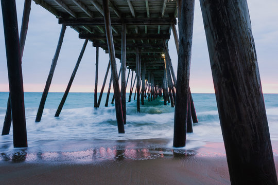 Sunrise under a beach fishing pier in the Outer Banks, NC. Long exposure image gives a softness to the surf. The light of the sunrise is reflected in the wet sand.