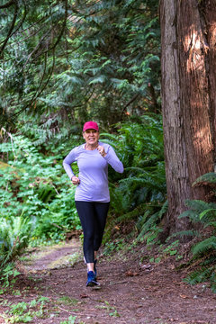 Baby Boomer Adult Woman With A Red Baseball Hat, Purple Shirt, Black Pants, And Blue Shoes Running On A Trail In The Woods
