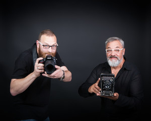 Fototapeta premium Two male photographers, young and elderly, with cameras - modern digital and vintage widescreen. On a black background in the studio.