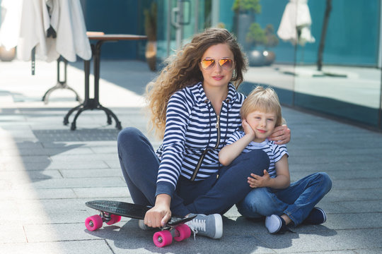 Young beautiful mother with her little cheerful son having fun and skating outdoors. Pretty cute kid with his mom together resting outside with skateboard. Happy family